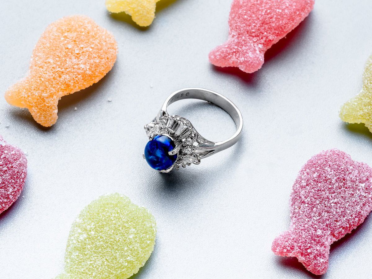 A silver ring with a round deep blue gemstone and small clear accent stones, styled on a light gray background with colorful sugar-dusted candy pieces around it.