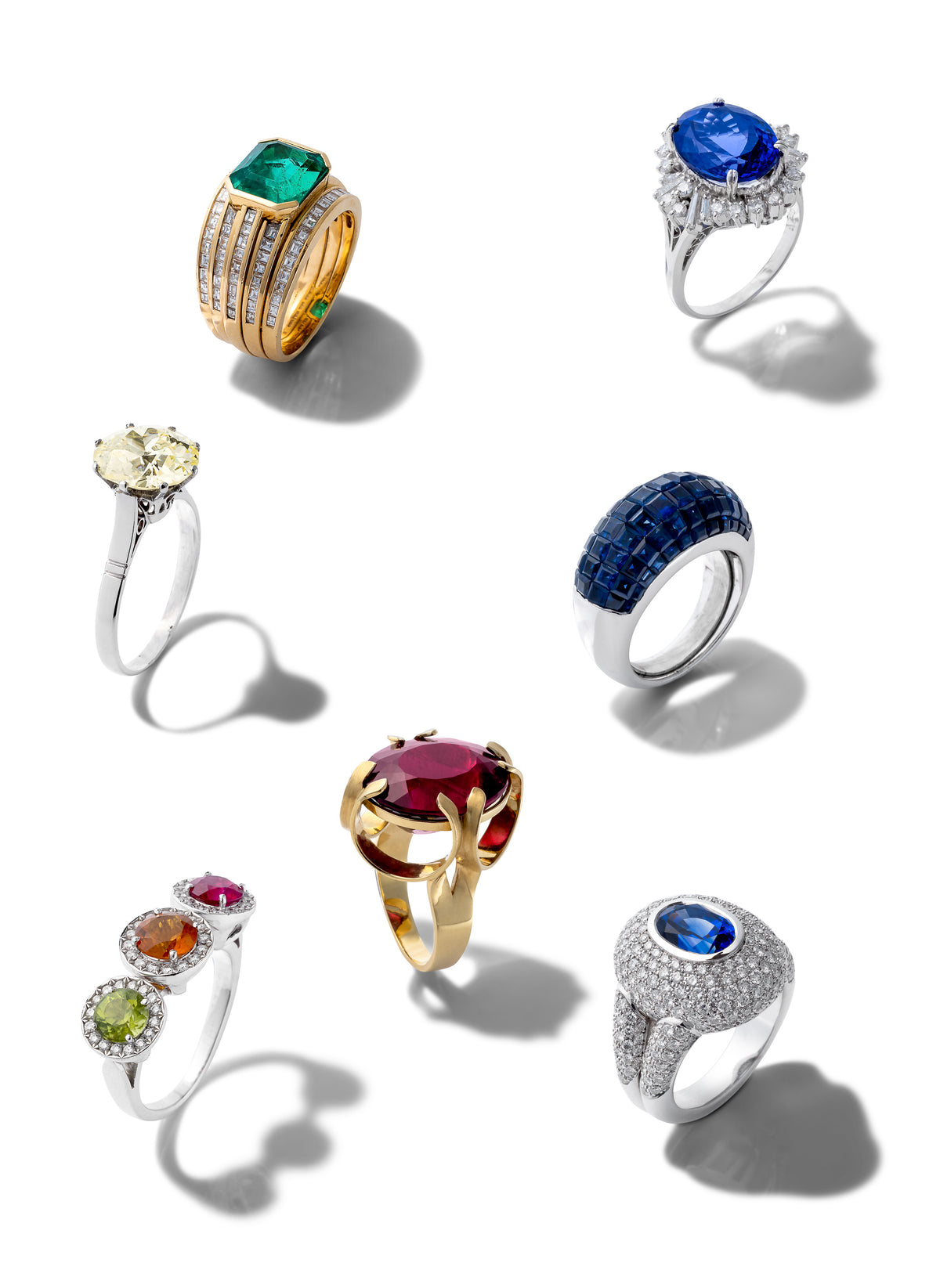 Seven distinct gemstone rings arranged on a clean white background with soft shadows. The collection includes yellow gold and silver settings with various colored gemstones such as emerald green, royal blue, sapphire blue, ruby red, yellow diamond, and round-cut clear stones.